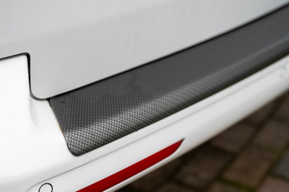 Close-up of a 2019 Volkswagen Transporter T28 Trendline TDI's rear bumper with a protective, textured surface. The white bumper features a black textured section and a red reflector at the bottom. The car is parked on a stone-paved driveway.