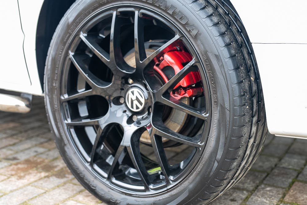 Close-up of a black car wheel with a prominent Volkswagen logo on the hub. The tire has visible tread patterns, and a red brake caliper is partially visible behind the rim. The 2019 Volkswagen Transporter T28 Trendline TDI is parked on a paved surface.