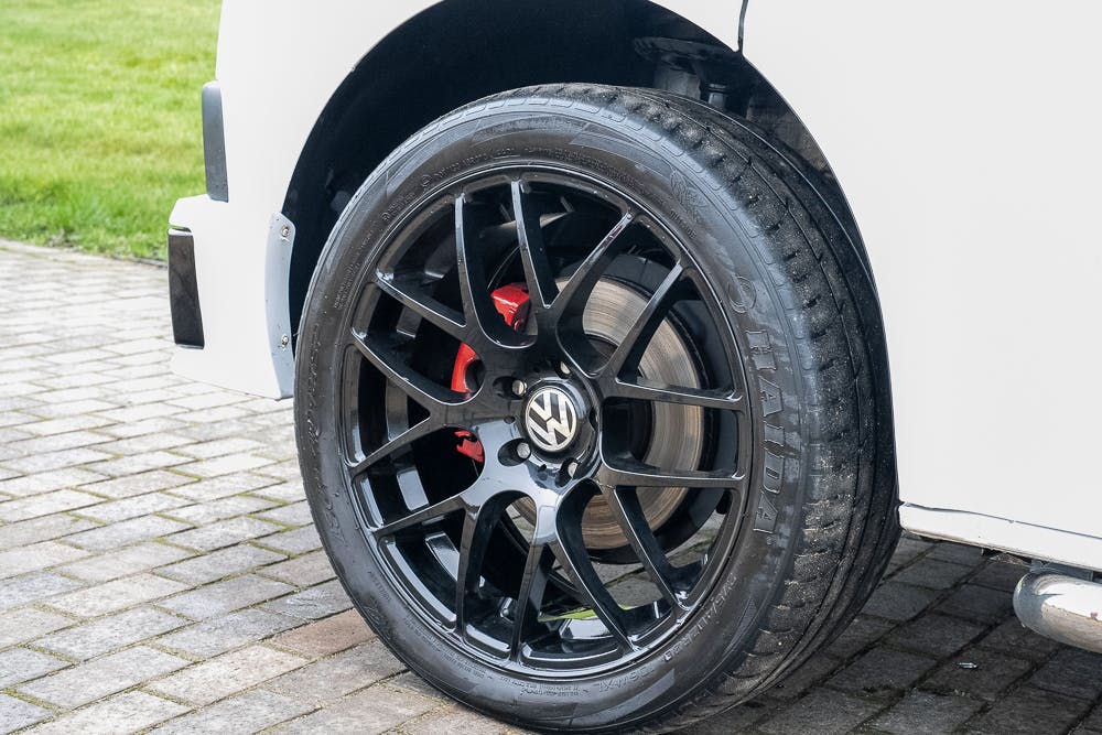 Close-up of a black alloy wheel with a Volkswagen logo in the center, attached to a white 2019 Volkswagen Transporter T28 Trendline TDI. The multi-spoke design reveals red brake calipers. The car is parked on a cobblestone driveway with grass in the background.