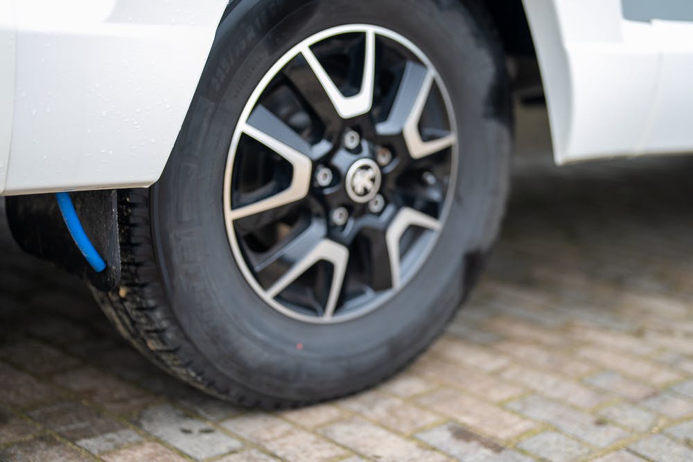 Close-up of a 2016 Roller Team Auto-Roller 707 Low Line's rear wheel on a paved surface. The black tire with visible treads is mounted on a black and silver alloy rim, while the vehicle's white body is partially visible in the image.