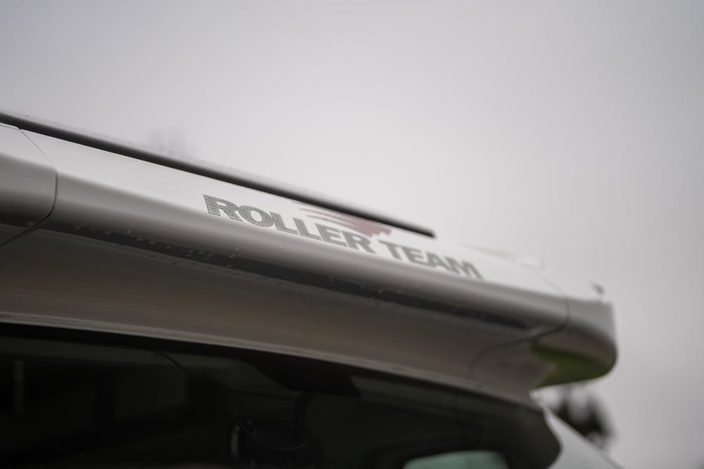 Close-up of the top section of a 2016 Roller Team Auto-Roller 707 Low Line RV with the brand name "Roller Team" visible. The background is out of focus and appears to show a gloomy, overcast sky.