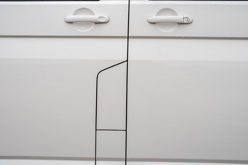 Close-up image of the side of a 2019 Volkswagen Transporter T28 Trendline TDI, displaying two door handles and part of the door panel. The image is minimalistic, showcasing the vehicle's clean lines and design details. There are visible water droplets on the surface, indicating recent rain.