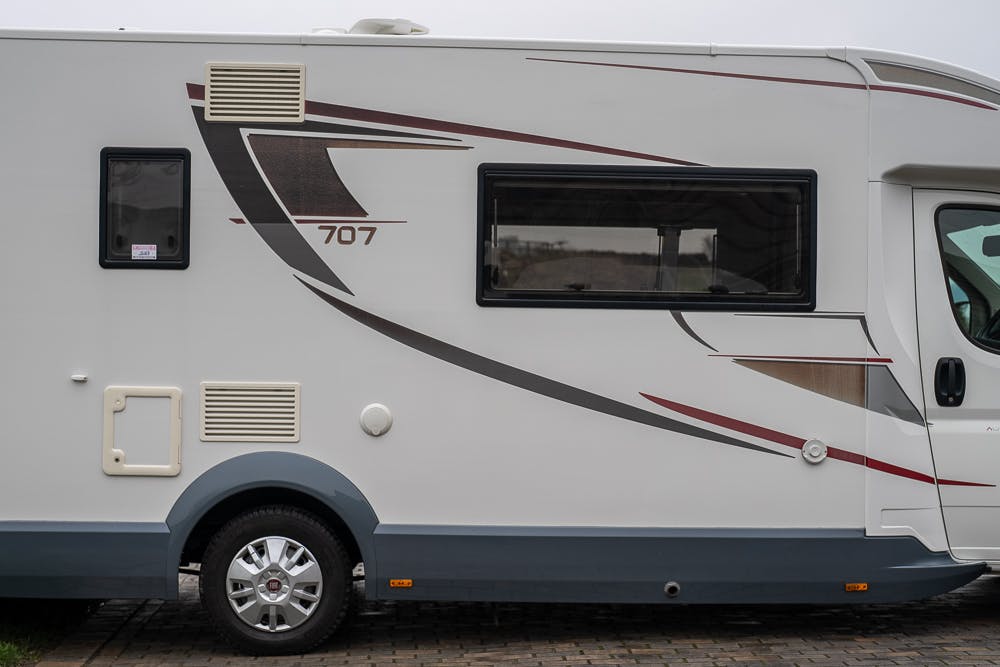 A side view of a white 2016 Roller Team Auto-Roller 707 Low Line RV, marked with the number 707. The RV has a large dark window and a pattern of grey and red lines. It is parked on a paved surface.