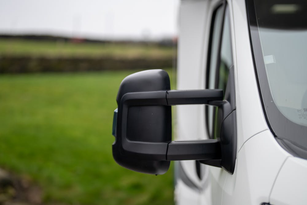 Close-up of a side mirror attached to a 2016 Roller Team Auto-Roller 707 Low Line white van. The background features a grassy field and a stone wall, slightly out of focus. The day appears overcast, as the lighting is soft and diffused.