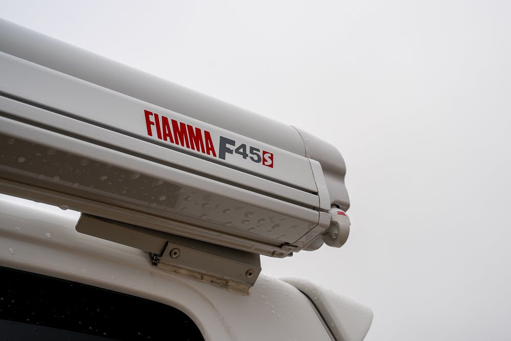 A close-up shot of a Fiamma F45S awning mounted on a 2019 Volkswagen Transporter T28 Trendline TDI. The awning is retracted, and there are droplets of water on its surface. The sky in the background is cloudy and overcast.