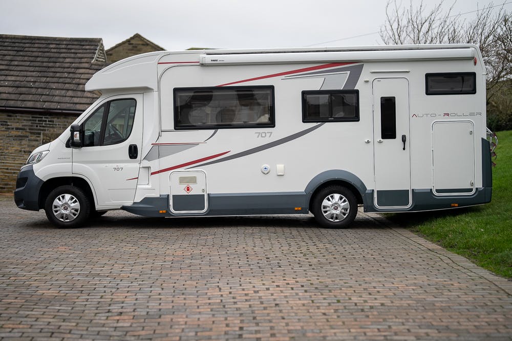 A 2016 Roller Team Auto-Roller 707 Low Line, white and gray motorhome, is parked on a brick driveway beside grass. The vehicle features multiple windows and compartments along its side. In the background, there's a part of a house with a slate roof and some bare tree branches.