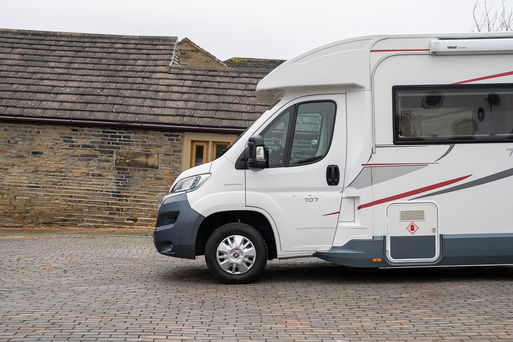 A 2016 Roller Team Auto-Roller 707 Low Line motorhome is parked on a brick-paved surface in front of a stone building with a tiled roof. The vehicle has a side door and window, and the front of the building features a small window and door.
