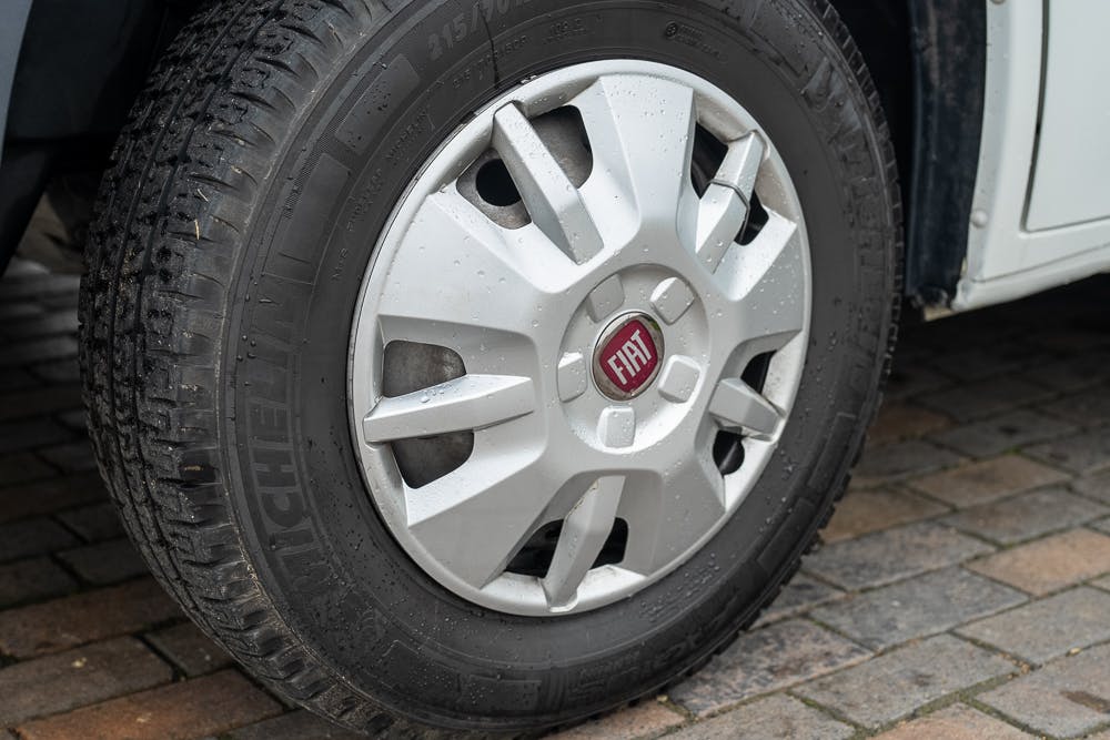 Close-up of a Fiat car wheel on a paved surface, parked next to a 2016 Roller Team Auto-Roller 707 Low Line. The tire brand is Michelin, and the hubcap features the Fiat logo in the center. The hubcap has a distinctive design with multiple spokes. The tire shows some dirt and light wear.