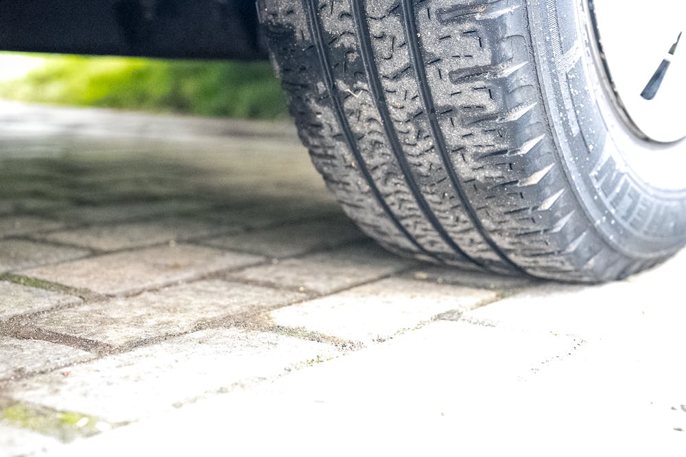 Close-up of a car tire with a patterned tread on the paved surface under the 2016 Roller Team Auto-Roller 707 Low Line. The ground consists of interlocking rectangular stones. The background shows some green grass and a shadow underneath the car.