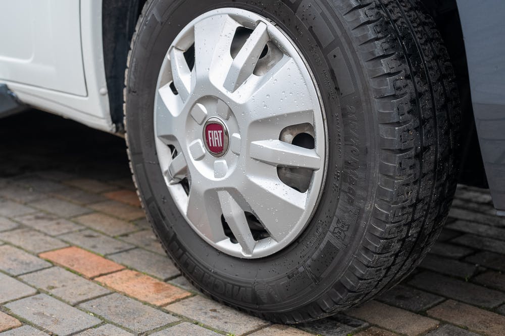 Close-up of a Fiat car wheel with water droplets on it. The tire, mounted on a silver alloy rim with the Fiat logo in the center, belongs to a 2016 Roller Team Auto-Roller 707 Low Line. The car is parked on a wet, paved surface with varying shades of gray and brown bricks.