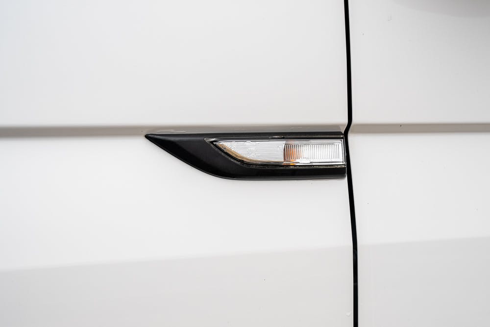 Close-up of a 2019 Volkswagen Transporter T28 Trendline TDI's side indicator light integrated into the side panel. The light has a clear lens with an orange bulb inside and is bordered with a black trim. The car's body is painted white.