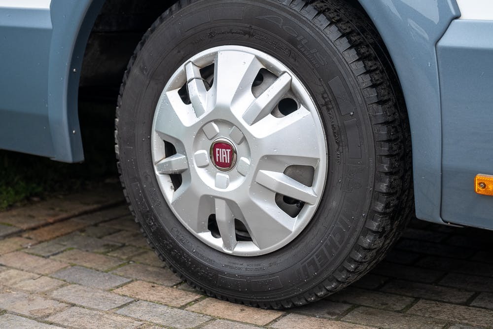 Close-up of a Fiat car tire with the Fiat logo in the center of the silver wheel. The tire is mounted on a grey vehicle, likely a 2016 Roller Team Auto-Roller 707 Low Line, and the image is taken from an angle that shows part of the wheel well and a small section of the ground below.