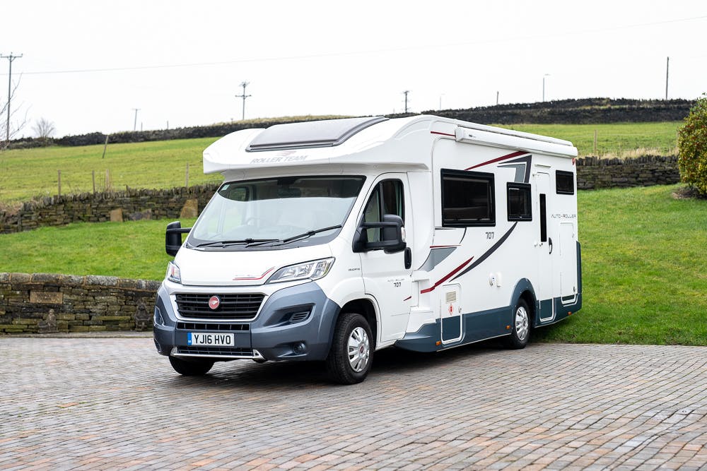 A 2016 Roller Team Auto-Roller 707 Low Line motorhome with the label "Roller Team" is parked on a paved driveway. The motorhome, featuring large side windows and a UK license plate YJ16 HVO, is set against a background of green grass and a stone wall.