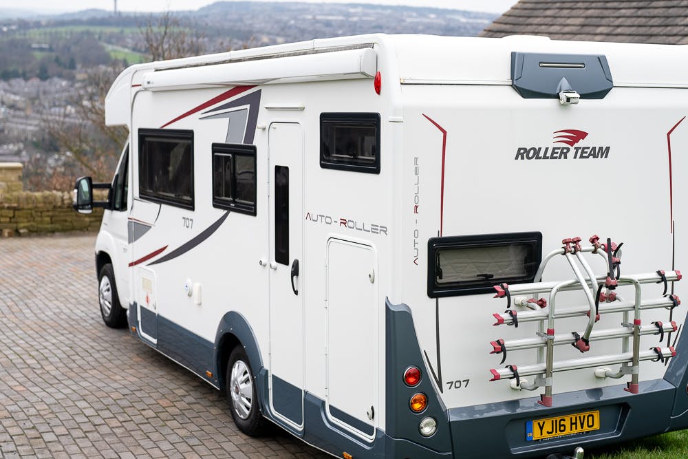 A 2016 Roller Team Auto-Roller 707 Low Line motorhome is parked on a driveway with a scenic view in the background. The white vehicle features a bike rack mounted on the rear, and its license plate reads YJ16 HVO. The ground is paved with bricks.