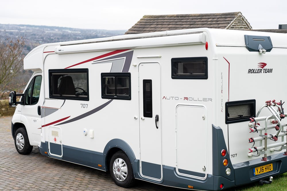 A white 2016 Roller Team Auto-Roller 707 Low Line motorhome with Roller Team branding is parked on a paved area. It features a rear bike rack, several windows, and grey and red graphics on the side. The background includes a cloudy sky and partial view of a structure.