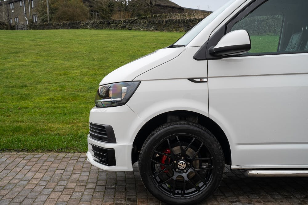 The image shows the front left side of a 2019 Volkswagen Transporter T28 Trendline TDI parked on a tiled pavement, adjacent to a grassy area. The focus is on the vehicle's front wheel and side mirror, with a background of trees and a stone wall.