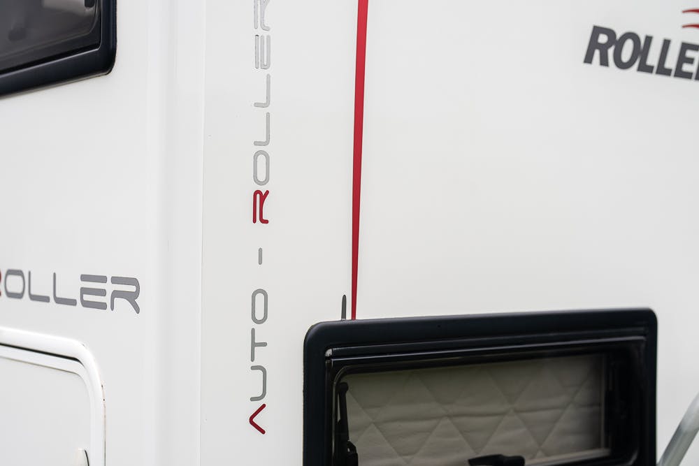 Close-up of the side of a 2016 Roller Team Auto-Roller 707 Low Line vehicle, showing the logo and part of a window with a quilted interior covering. The text "AUTO-ROLLER" is visible in red and gray lettering on the white surface.