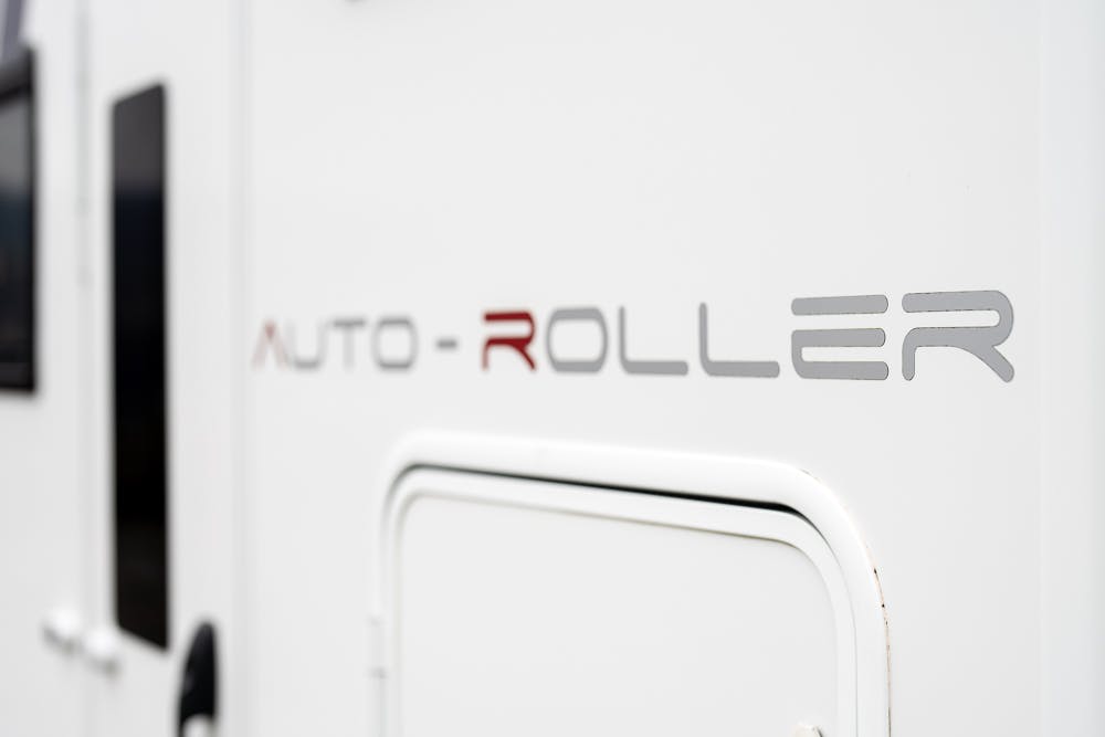 Close-up of a white 2016 Roller Team Auto-Roller 707 Low Line with "Auto-Roller" written on the side in silver and red letters. The image also shows part of a black window and a white door.