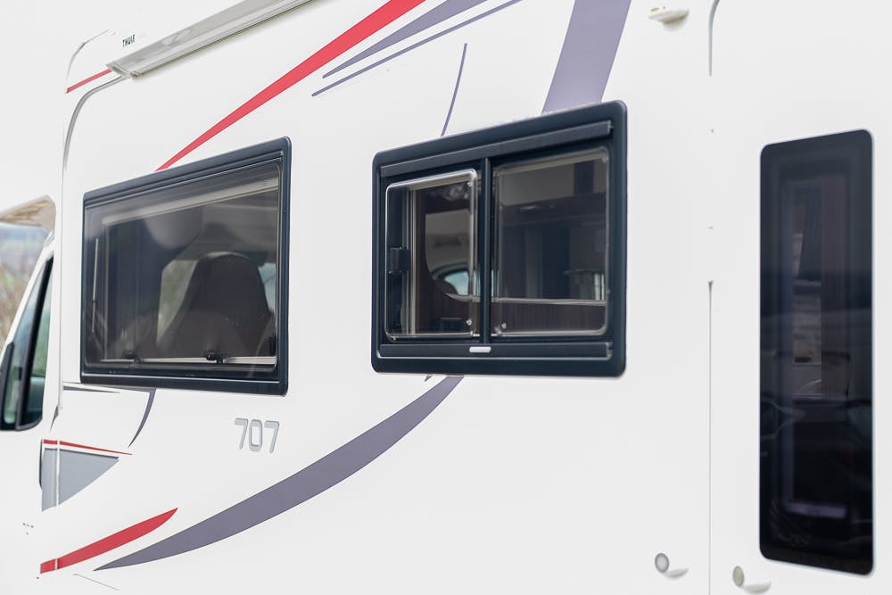 Close-up of the side of a 2016 Roller Team Auto-Roller 707 Low Line RV, with the number 707 printed on it. The RV has two black-framed windows, with the larger one to the left and the smaller one to the right. The side is accented with red and gray stripes.