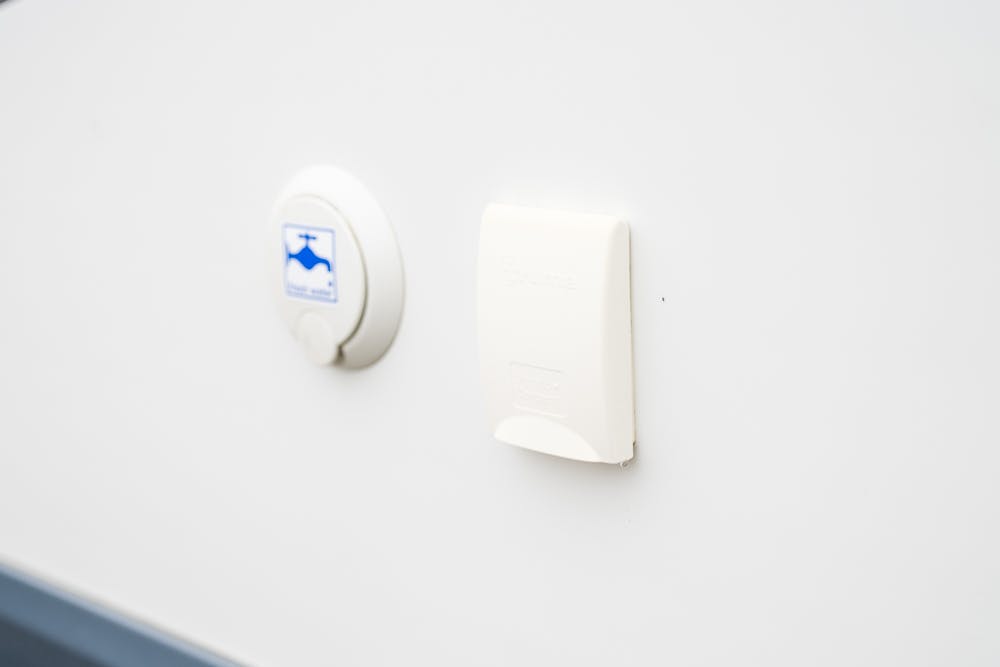 A white wall features two covered outlets. The outlet on the left has a round, circular cover with a blue bird logo reminiscent of the 2016 Roller Team Auto-Roller 707 Low Line design. The outlet on the right has a rectangular cover with "Bayonet" imprinted on it. Both covers are closed.