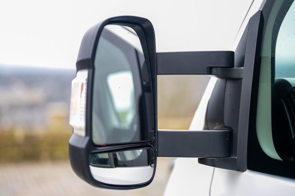 Close-up view of a side mirror attached to a 2016 Roller Team Auto-Roller 707 Low Line, featuring an extended arm and an integrated turn signal light. The mirror has two sections for better visibility. The background is blurred.