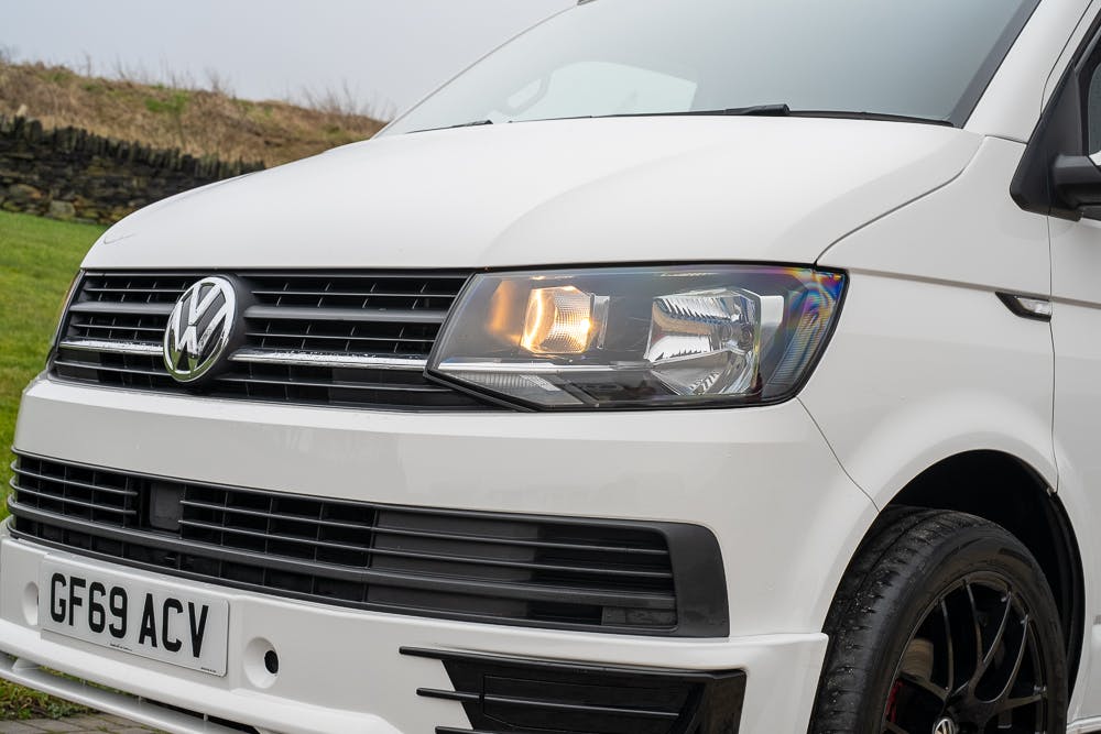 Close-up view of the front end of a white 2019 Volkswagen Transporter T28 Trendline TDI with license plate GF69 ACV. The car's right headlight is on, and the vehicle is parked outdoors on a concrete surface with a grassy backdrop.