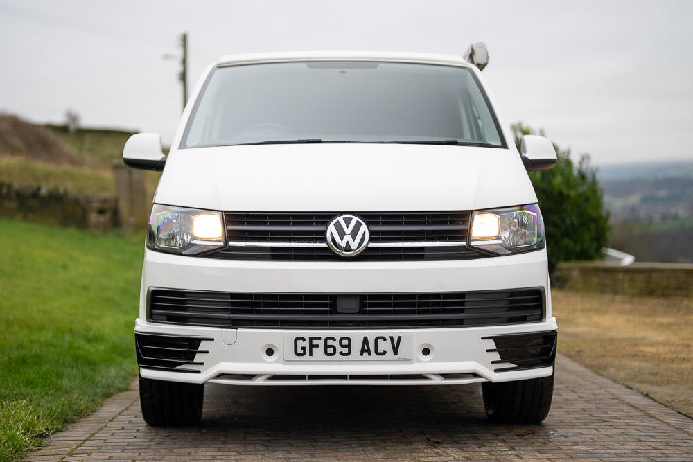 Front view of a white 2019 Volkswagen Transporter T28 Trendline TDI van with license plate GF69 ACV, parked on a cobblestone driveway. The headlights are illuminated, casting a warm glow. The background features greenery and a cloudy sky.