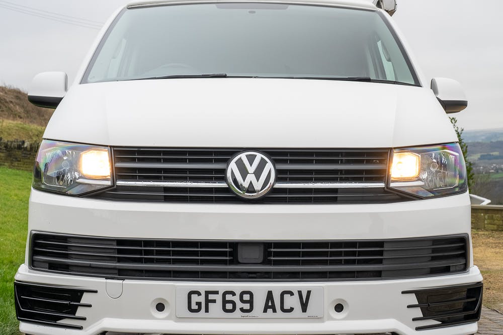 A white 2019 Volkswagen Transporter T28 Trendline TDI van is shown from the front with headlights on. The license plate reads "GF69 ACV." The background features a grassy area and trees under a cloudy sky.
