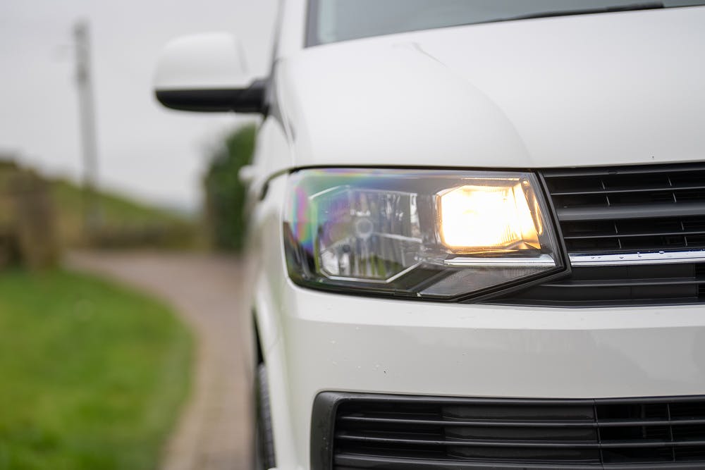 Close-up view of the front right headlight of a 2019 Volkswagen Transporter T28 Trendline TDI with the headlight turned on. There is a road and some greenery visible in the background on the left side. The image captures a partial view of the vehicle.