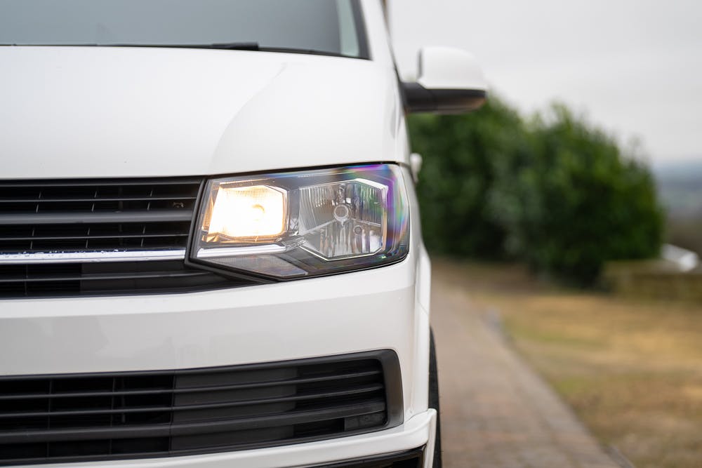 Close-up of the front right headlight of a white 2019 Volkswagen Transporter T28 Trendline TDI with the right side headlight illuminated. The background is slightly blurred, showcasing a paved pathway and green bushes, suggesting the image was taken on an overcast day.
