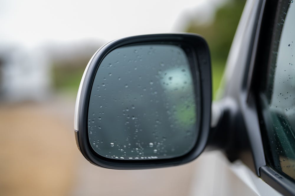 Close-up of a 2019 Volkswagen Transporter T28 Trendline TDI's side mirror with water droplets on the glass. The background is blurred, indicating an outdoor setting with wet weather conditions. The mirror housing is partially visible, showcasing a typical side mirror design and attachment to the vehicle.