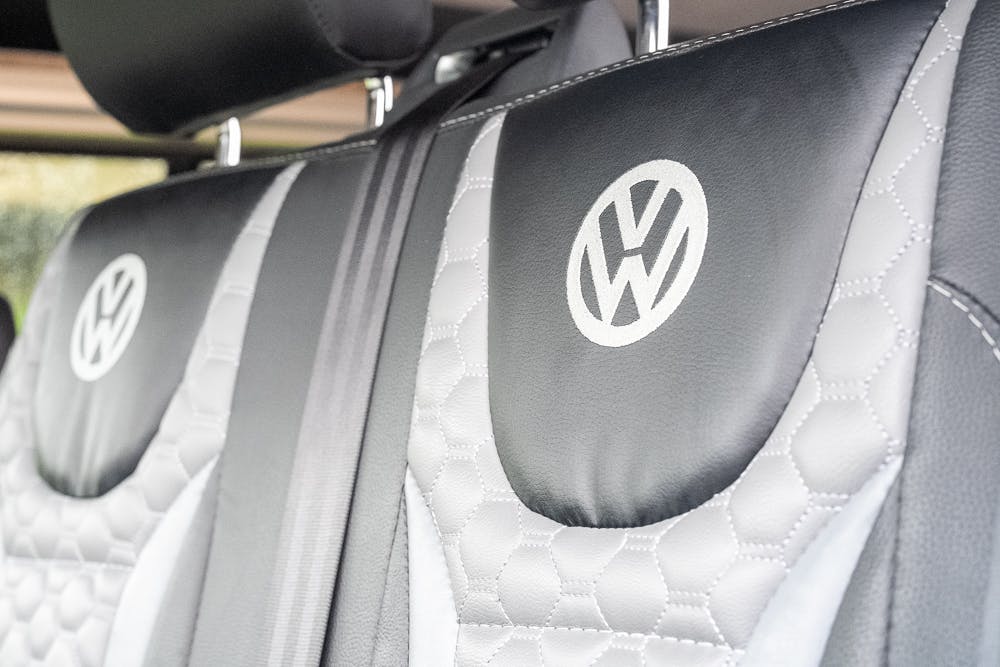 Close-up view of two car seats with black and gray leather upholstery, featuring the Volkswagen logo stitched at the top of each seat in the 2019 Volkswagen Transporter T28 Trendline TDI. The leather has a hexagonal pattern along the sides. The headrests are visible at the top of the image.