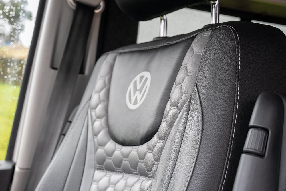 Close-up of a car seat with a VW (Volkswagen) emblem stitched on the backrest. The seat of this 2019 Volkswagen Transporter T28 Trendline TDI features a two-tone design with black and gray leather, including a honeycomb pattern in the gray areas. The background shows part of the car interior and a window with raindrops.