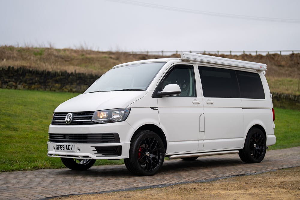 A 2019 Volkswagen Transporter T28 Trendline TDI van with black wheels is parked on a paved area near a grassy field. Sporting a UK license plate "GF69 ACV" and featuring a roof awning, the white van is set against a backdrop of stone wall and cloudy sky.