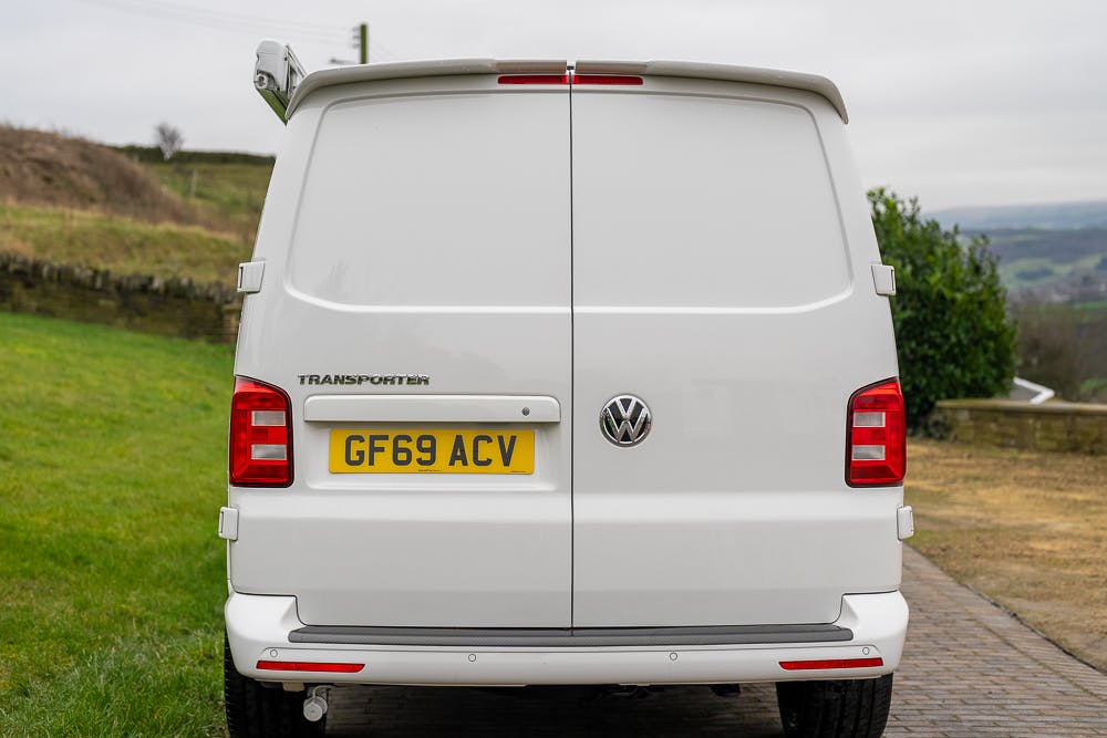 A 2019 Volkswagen Transporter T28 Trendline TDI is parked on a paved driveway. The rear view shows the vehicle's license plate "GF69 ACV" and the backdoors are closed. The background features a grassy hillside and cloudy sky.