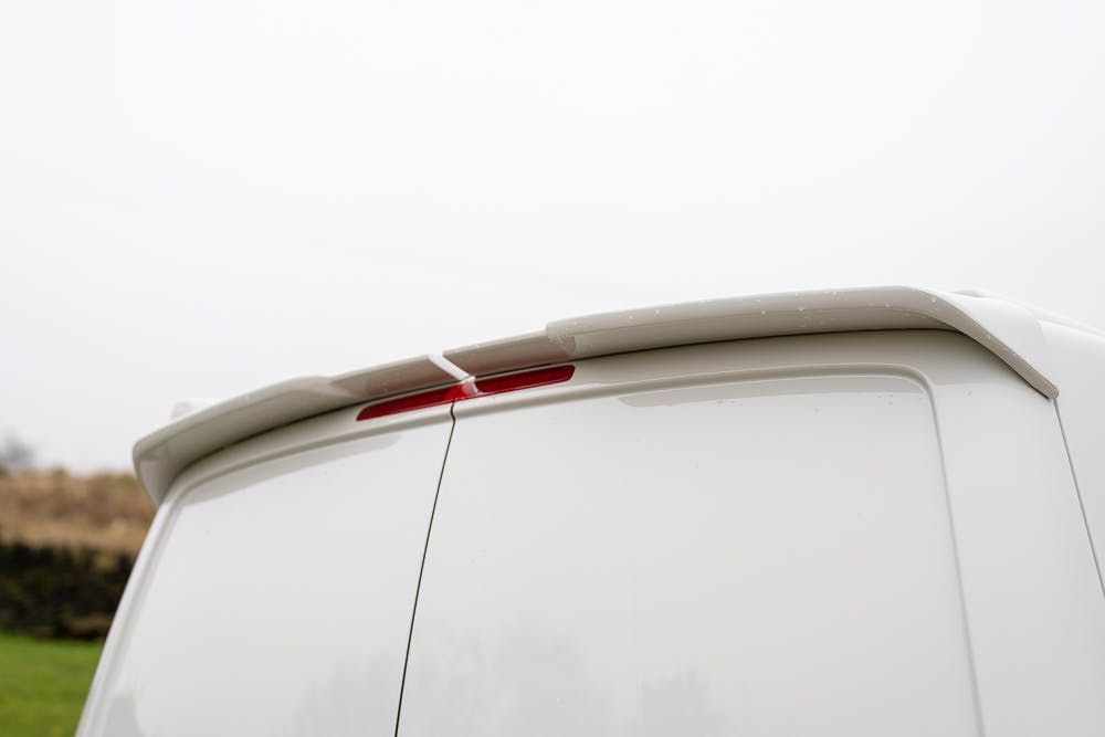 The image shows the rear portion of a white 2019 Volkswagen Transporter T28 Trendline TDI, focusing on the roofline and rear door. A red brake light is visible just below the roof spoiler. The background is blurred and appears to be an outdoor setting.