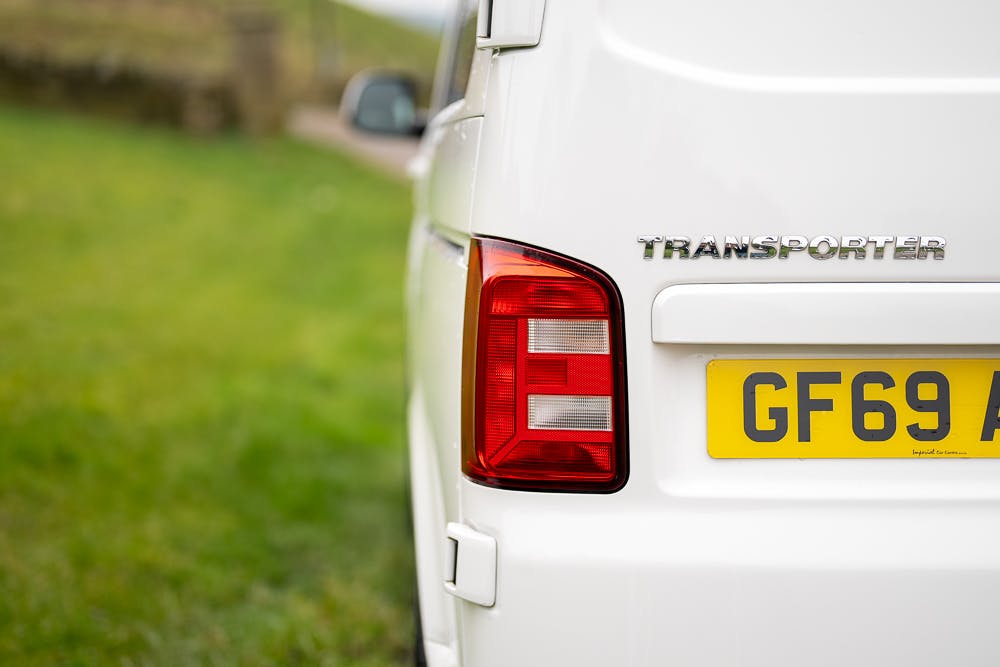 Close-up view of the rear left corner of a 2019 Volkswagen Transporter T28 Trendline TDI in white, featuring "Transporter" on the back and a yellow license plate reading "GF69 A". The vehicle is parked on a grassy area, with a blurry background of more grass and a road.