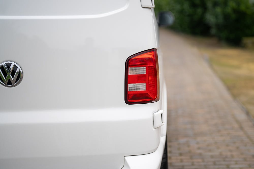 Close-up of the rear left taillight of a 2019 Volkswagen Transporter T28 Trendline TDI, featuring a red and white light assembly. The white van, parked on a paved driveway with some greenery in the background, displays part of its emblem on the left side.
