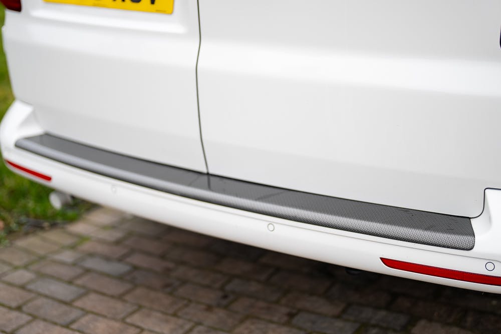 The image shows the rear lower section of a white 2019 Volkswagen Transporter T28 Trendline TDI with part of the license plate visible at the top left corner. The bumper has a strip of dark material, likely for protection, and red reflectors are positioned near the sides. The car is parked on a brick surface.