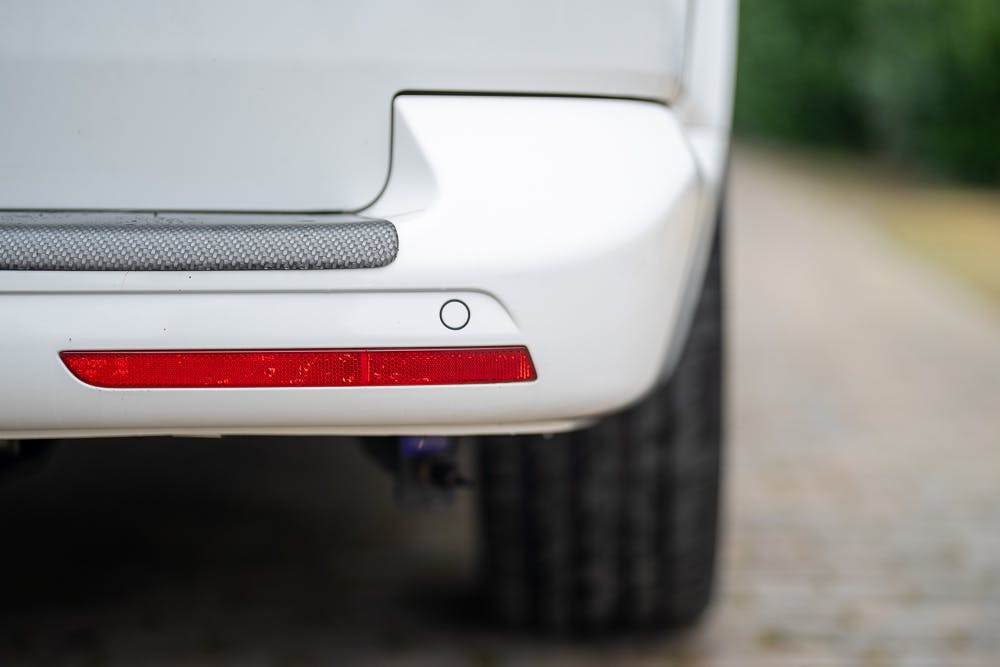Close-up image of a 2019 Volkswagen Transporter T28 Trendline TDI's rear bumper showing a red reflector and a sensor. The car is parked on a cobblestone surface with some greenery blurred in the background.