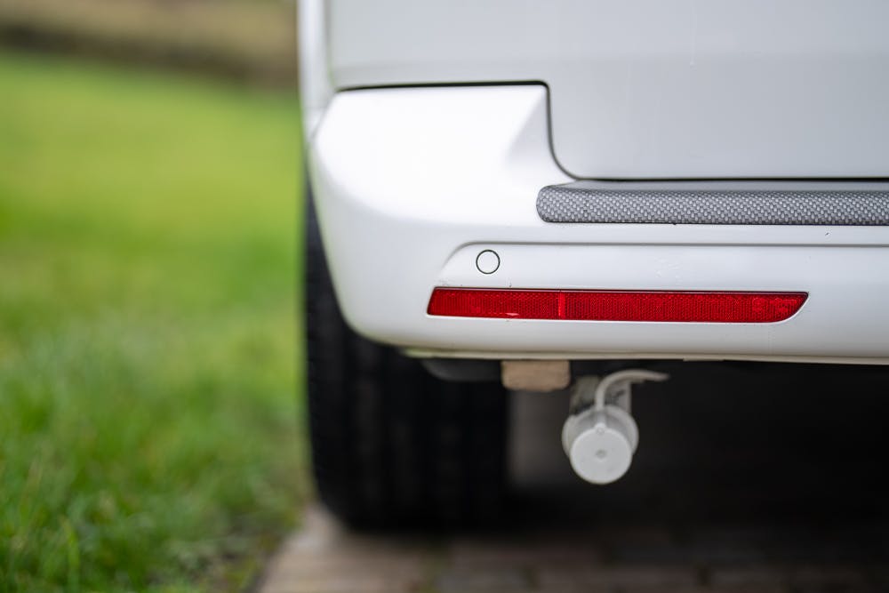 Close-up of the rear bumper of a 2019 Volkswagen Transporter T28 Trendline TDI, showing a red reflector and a tow hook cap. The background is blurred, with a grassy area and a paved surface partially visible.