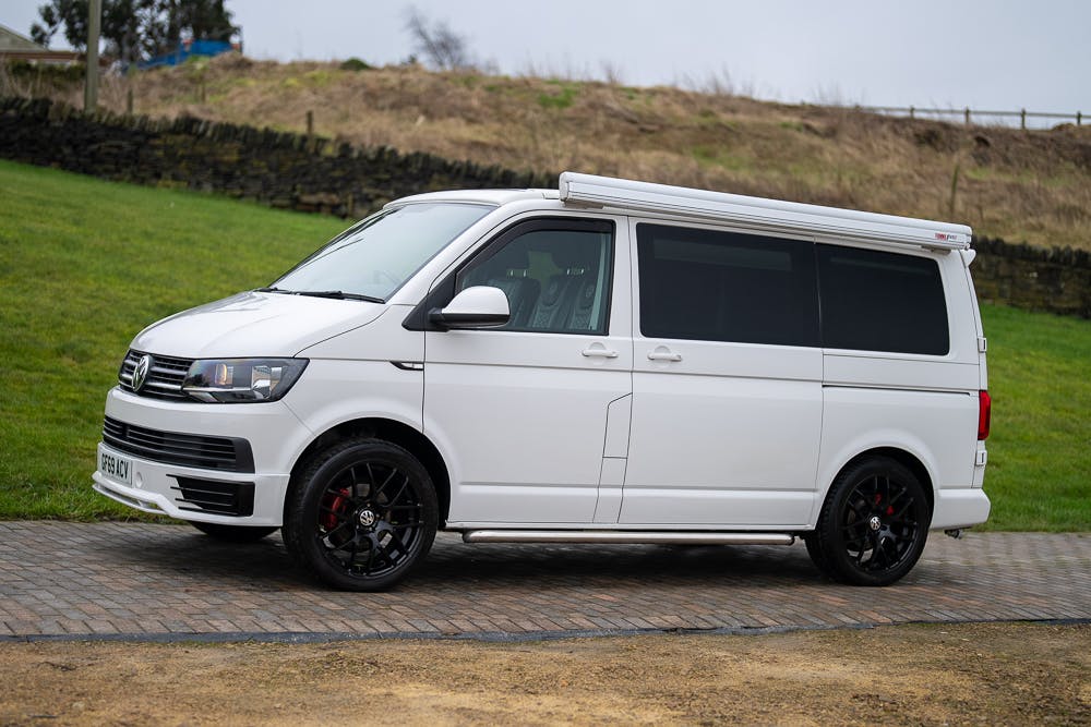 A 2019 Volkswagen Transporter T28 Trendline TDI camper van is parked on a paved area. The vehicle, featuring black wheels, tinted windows, and an extended roof with a built-in awning, sits against a backdrop of a grassy hill and stone wall. The sky is overcast.