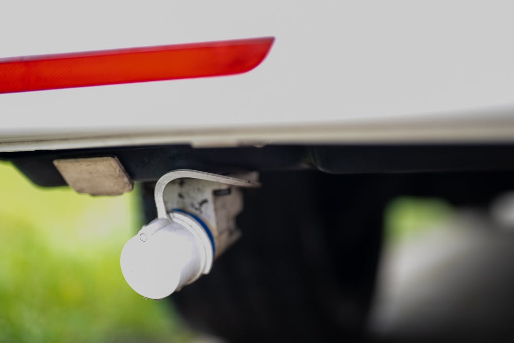 Close-up of a trailer hitch with a mounted electrical connector on the rear of a 2019 Volkswagen Transporter T28 Trendline TDI. The red reflector on the bumper is partially visible, and the background shows a blurred green lawn and driveway.