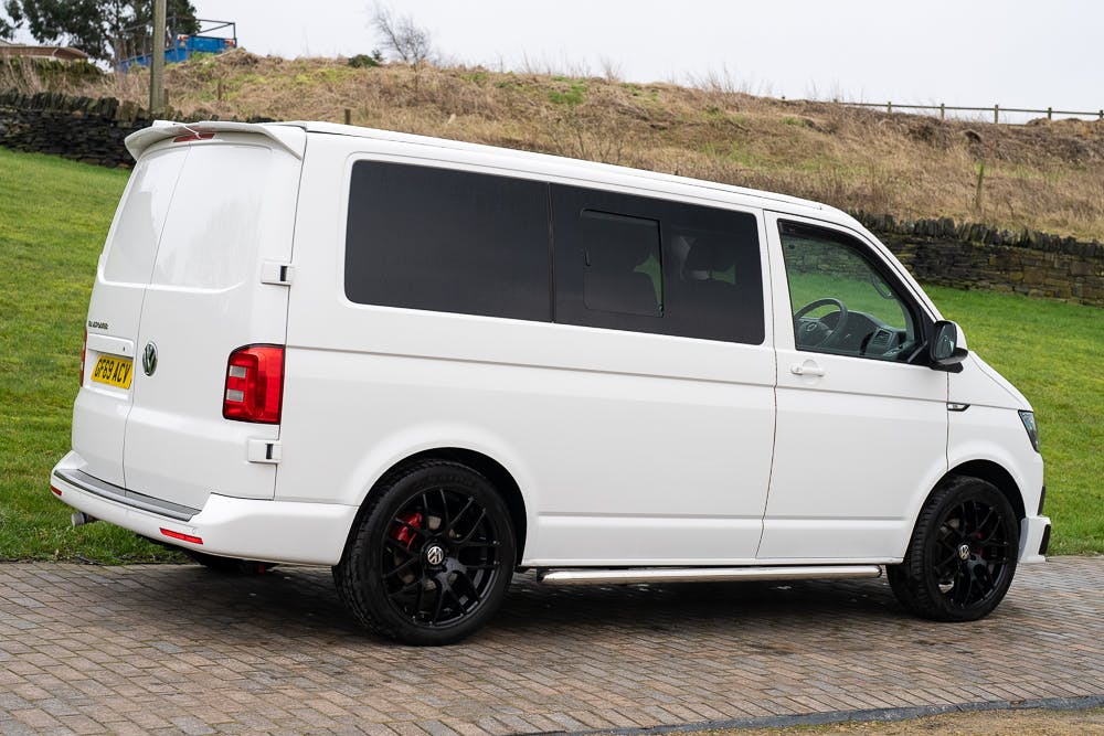 A 2019 Volkswagen Transporter T28 Trendline TDI with tinted windows is parked on a sloped, paved surface. The white van sports black alloy wheels and a UK license plate. In the background, a grassy area, stone wall, and some bare trees complete the scene.