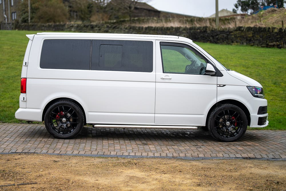 A white 2019 Volkswagen Transporter T28 Trendline TDI is parked on a paved surface with a grassy area and stone wall in the background. The van features dark-tinted windows and black alloy wheels, set against the backdrop of a serene rural landscape.