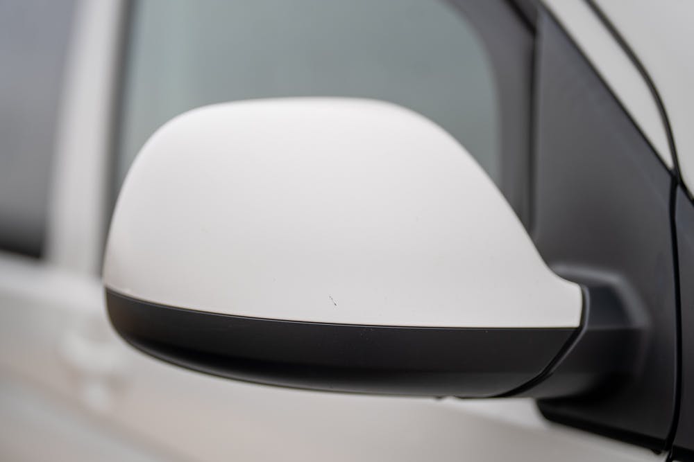 Close-up of the side mirror on a 2019 Volkswagen Transporter T28 Trendline TDI. The mirror housing is partially white with a black section at the bottom. In the background, part of the car's window and frame is visible, with focus on the side mirror.
