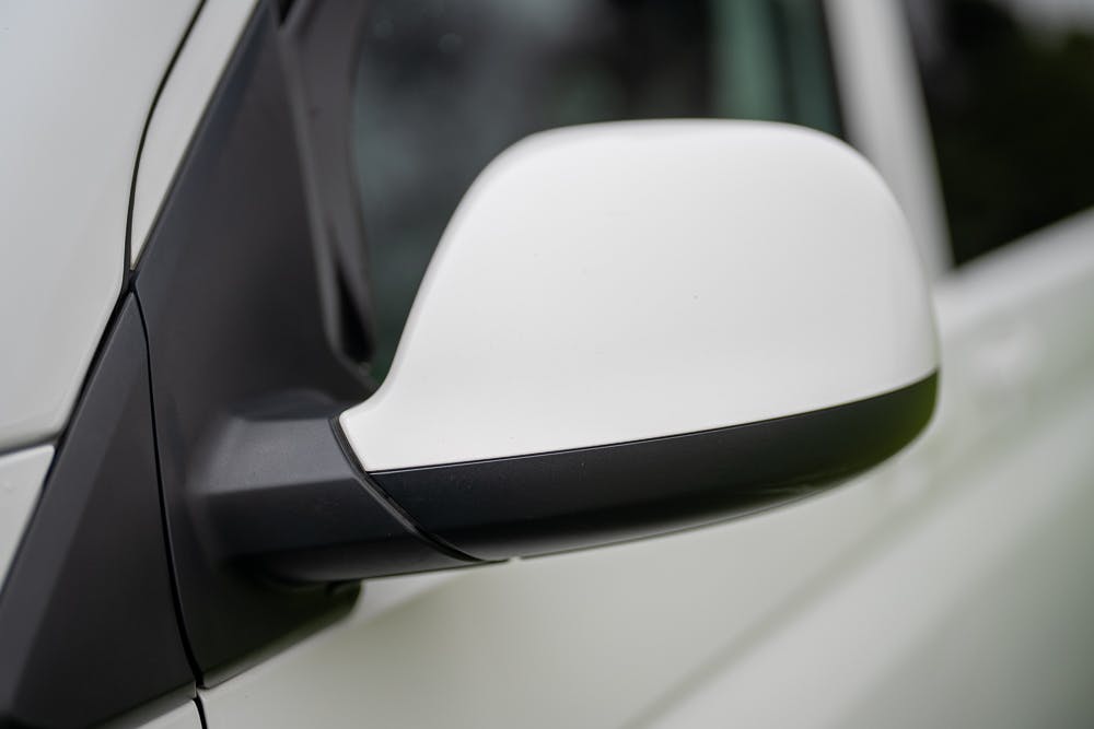 Close-up of a 2019 Volkswagen Transporter T28 Trendline TDI's white side mirror with a black casing. The background is blurred, highlighting the focus on the side mirror. The car appears to be clean and in good condition.