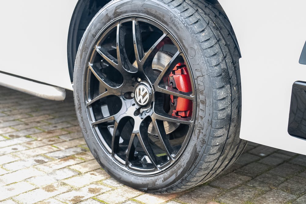 Close-up view of a black alloy wheel with a visible Volkswagen logo on the center cap, and a red brake caliper behind it, on a 2019 Volkswagen Transporter T28 Trendline TDI parked on a cobblestone surface.