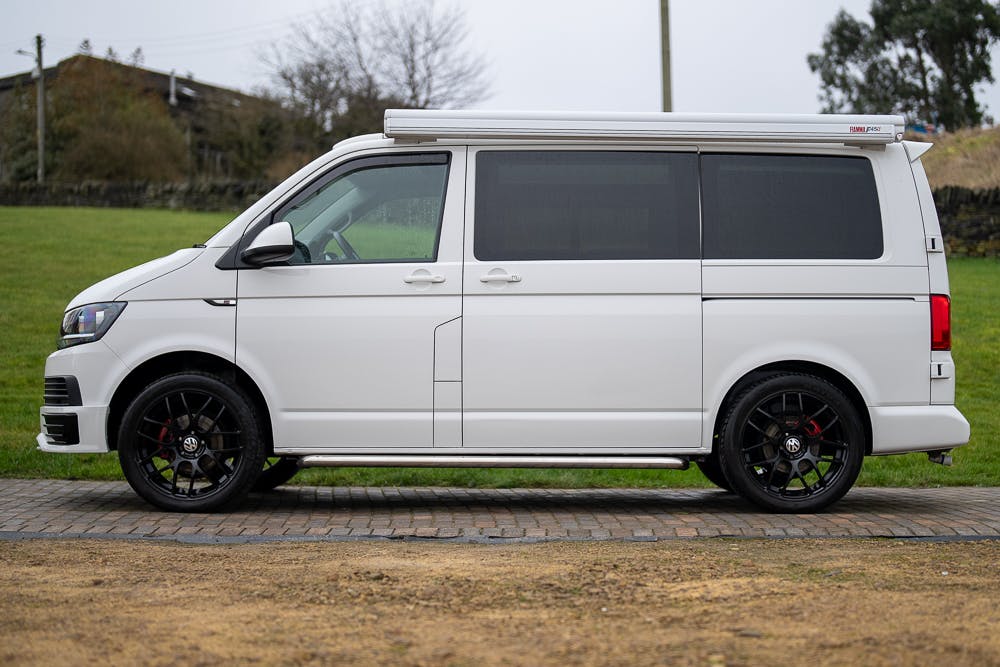 A 2019 Volkswagen Transporter T28 Trendline TDI camper van is parked outdoors on a paved surface with a grassy area and trees in the background. The van features tinted windows, black wheels, and a roof rack.