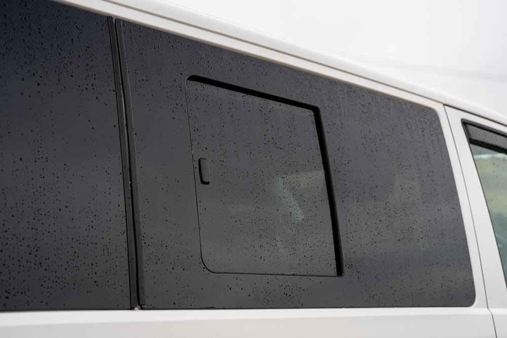 A close-up view of a 2019 Volkswagen Transporter T28 Trendline TDI's side window, adorned with raindrops. The window features a small rectangular vent with a handle. The scene is likely outdoors on a rainy day.